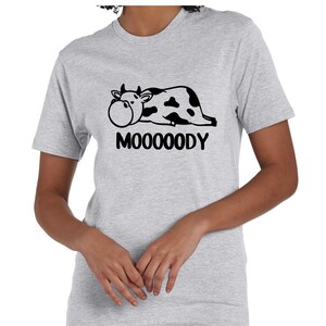 May include: Light gray t-shirt featuring a black and white cow graphic with the word "MOOOOODY" below. The cow is lying down with a large, round face. The shirt has short sleeves and a crew neck.