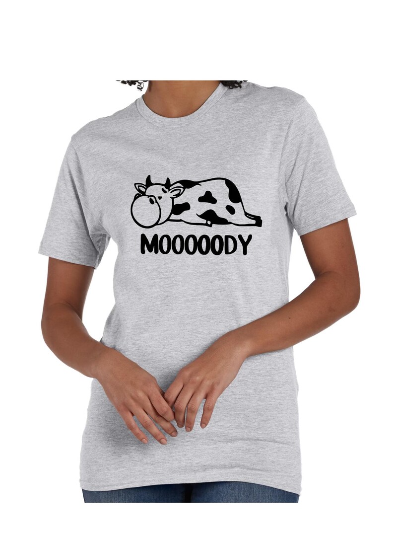 May include: Light gray t-shirt featuring a black and white cow graphic with the word "MOOOOODY" below. The cow is lying down with a large, round face. The shirt has short sleeves and a crew neck.