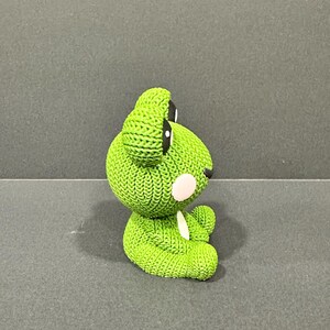 May include: A green knitted frog figurine with black eyes and a white belly. The frog has a textured, bumpy surface and is sitting on a gray surface. The frog's cheeks are a light pink color.