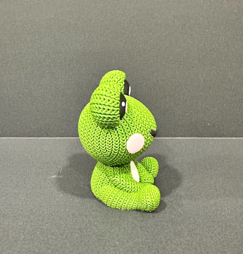 May include: A green knitted frog figurine with black eyes and a white belly. The frog has a textured, bumpy surface and is sitting on a gray surface. The frog's cheeks are a light pink color.