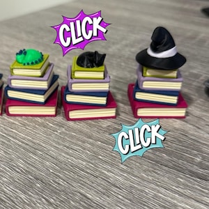 May include: Miniature stacks of books topped with a green creature, a black cat, and a witch's hat. The books are in shades of red, purple, and blue. The word "CLICK" appears in a comic book style speech bubble.