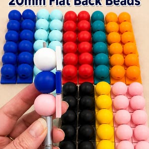 May include: Rows of colorful 20mm flat back beads in various colors, including blue, red, orange, yellow, and pink. The beads are arranged on rectangular bases. The image also shows a pen with beads attached to it.