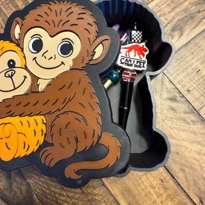 May include: A 3D-printed monkey-shaped box with a lid featuring a smiling monkey hugging another monkey. The box is black with brown and orange accents. Inside the box are various pens and a sticker that says "CAN I PET THAT DOG?"