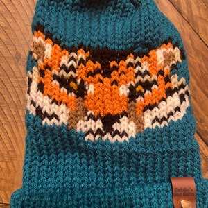 Può includere: Berretto lavorato a maglia color teal con un disegno di volpe arancione, bianca, nera e marrone. Il cappello ha un pompon in finta pelliccia bianca e una piccola etichetta in pelle con "Goldie's Girl Gifts".