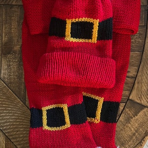 May include: Red knitted Santa hat with a white pom pom and a black and gold belt design. The hat is paired with a matching red scarf with a black and gold belt design and white fur trim.