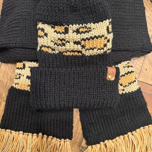 May include: Black knit hat with a brown and tan geometric pattern and a faux fur pom pom. The hat is paired with a matching black knit scarf with brown and tan fringe.