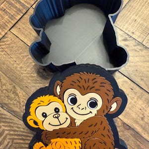 May include: A dark blue monkey-shaped cookie cutter with a gray interior sits above a monkey-themed sticker. The sticker features two cartoon monkeys hugging, one brown and one yellow, on a black background.