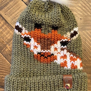May include: Green knitted beanie with a white pom pom on top. The beanie features a brown, orange and white fox design on the front. The beanie has a brown leather tag with a white swirl design.