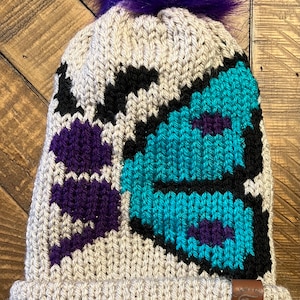 May include: A beige knitted beanie with a purple pom pom and a colorful butterfly design. The butterfly is made of teal, purple, and black yarn. The beanie has a small brown leather tag with a heart on it.