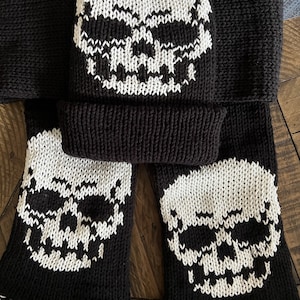 Puede incluir: Conjunto de gorro y bufanda de punto negro con diseños de calaveras blancas. El gorro tiene un puño abatible y la bufanda tiene flecos.