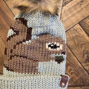 May include: A knitted beanie in shades of blue and brown, topped with a fluffy, light brown pom-pom. The hat features a geometric design and a small leather tag with a logo.