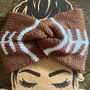 May include: Brown and white knitted headband with a football design. The headband is tied in a knot at the top.