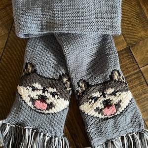 May include: A gray knitted scarf with a white and brown husky dog face design on each end. The scarf has long gray and white fringe on each end.