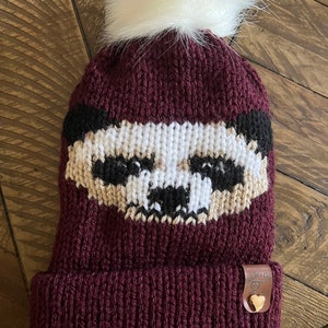 May include: A burgundy knitted beanie with a white pom pom on top. The beanie has a black and white badger face design on the front. The beanie has a brown leather tag with a gold heart on it.