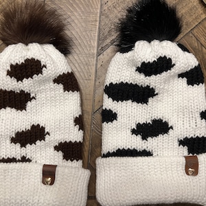 Puede incluir: Dos gorros de punto con estampado de vaca y pompones de piel sintética. Un gorro tiene manchas de vaca marrones y el otro tiene manchas de vaca negras. Ambos gorros tienen un puño blanco.