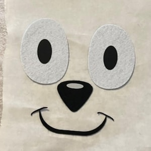 May include: A white felt applique of a smiling face with black eyes, nose, and mouth. The applique is on a white background.