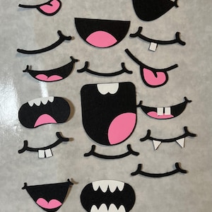 May include: A collection of black felt mouth and tongue cutouts with pink and white accents. The set includes various shapes and sizes of mouths, tongues, and teeth, suitable for crafting or educational purposes. The cutouts are arranged on a light gray surface.