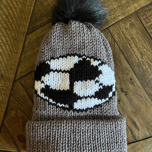 May include: A grey knitted beanie with a black and white striped design and a black faux fur pom pom on top.