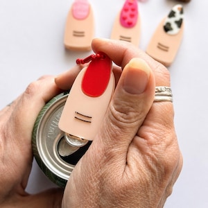 Könnte beinhalten: Ein beigefarbener Dosenöffner aus Kunststoff in Fingerform mit einem leuchtend rot lackierten Fingernagel und zwei horizontalen schwarzen Linien, die ein Fingergelenk darstellen. Das Werkzeug hat eine rote Kugelkette am oberen Ende.