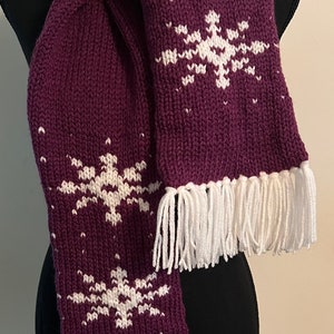 Könnte beinhalten: Ein lila gestrickter Schal mit weißen Schneeflockenmustern und weißen Fransen an den Enden.