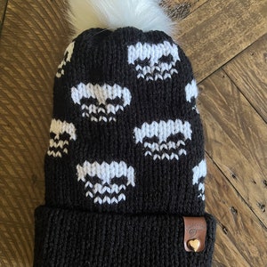 May include: Black knit beanie with a white pom pom and a white skull pattern. The beanie has a brown leather tag with a heart and the text "Charlie's Girl".