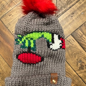 May include: Gray knitted beanie with a red pom pom and a green, white, and red embroidered design. The beanie has a brown leather tag with a gold heart.