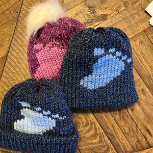 May include: Three knitted hats with pom poms. The hats are blue, pink, and dark blue with a white pom pom on top. Each hat has a blue and white design of a lake.