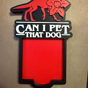 May include: A red and black sign with the white text "CAN I PET THAT DOG". The sign features a red dog silhouette and a flower. The background is red.