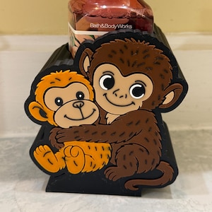 Könnte beinhalten: Ein schwarzer dekorativer Halter mit zwei Cartoon-Affen in Braun- und Orangetönen. Die Affen umarmen sich. Im Hintergrund befindet sich eine Flasche von Bath & Body Works.