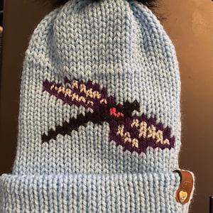 May include: A light blue knitted beanie with a black faux fur pom pom on top. The beanie has a black and purple embroidered dragonfly design on the front. The beanie has a brown leather patch with a silver heart on the side.