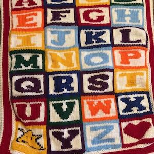 May include: A knitted alphabet blanket with colorful squares featuring each letter of the alphabet. The blanket has a white background with a red border.
