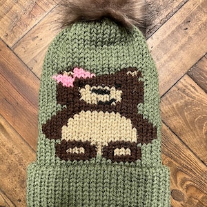 May include: Green knitted beanie with a brown bear design and a brown faux fur pom pom on top.
