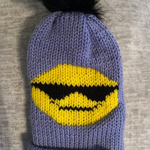 Peut inclure: Bonnet en tricot violet avec un pompon noir sur le dessus et un smiley jaune avec des lunettes de soleil noires sur le devant. Le bonnet a une étiquette en cuir marron avec un cœur.