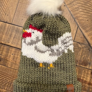 Puede incluir: Un gorro de punto verde con un pompón blanco y un diseño de pollo tejido en blanco y gris. El gorro tiene una etiqueta de cuero marrón con el texto "Good Vibes Only" en ella.