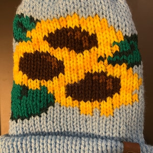May include: A light blue knitted beanie with a pom pom on top. The beanie features a design of yellow sunflowers with brown centres and green leaves. The beanie has a brown leather tag with a heart logo that says "Grace & Lace".