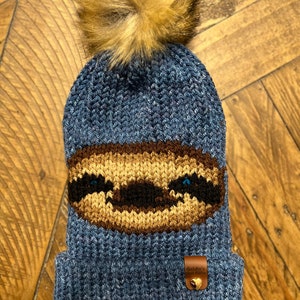 Puede incluir: Gorro azul de punto con una cara de perezoso marrón y un pompón de piel sintética marrón en la parte superior. El gorro tiene una etiqueta de cuero marrón con el texto "Goldie's Good Goods" y un corazón dorado.
