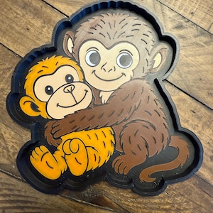 May include: A brown and yellow monkey-themed wall hanging. The image features two cartoon monkeys hugging each other. The monkeys are set against a black background. The wall hanging is made of a textured material.