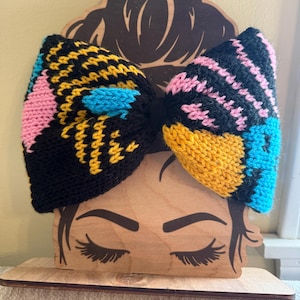 Peut inclure: Un grand bandeau en forme de nœud tricoté coloré en noir, jaune, rose et bleu. Le nœud est présenté sur un présentoir en bois avec un visage de femme. L'accessoire est vif.