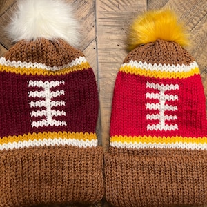 May include: Two knitted winter hats with pom poms. The hats are brown, red, white, and gold. The hats have a football design on the front.