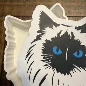 May include: A white cat-shaped trinket box with a black face and blue eyes. The box is open, revealing a white interior. The cat's face is detailed with black fur markings and whiskers. The box has a scalloped edge.