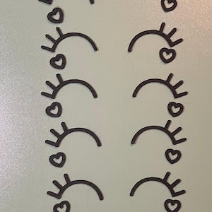 May include: Ten black paper cutouts of a stylized eye with eyelashes and a heart. The eyes are arranged in two columns of five.