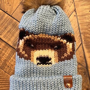 May include: A blue knitted beanie with a brown bear face design and a brown faux fur pom pom on top. The beanie has a leather patch with the text "Cozy Creations" and a small heart.