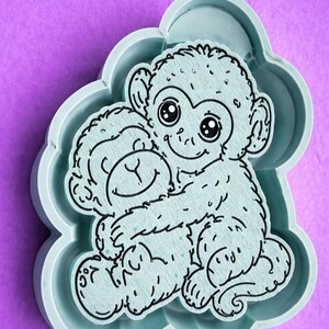 May include: A light blue monkey-shaped mold with a cartoon illustration of two monkeys hugging. The mold has a cloud-like shape and is set against a purple background. The monkeys have large eyes and are smiling.