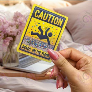 May include: Yellow caution sign with a stick figure falling on a bed of colorful beads. The sign reads "CAUTION BEADS ON THE FLOOR".