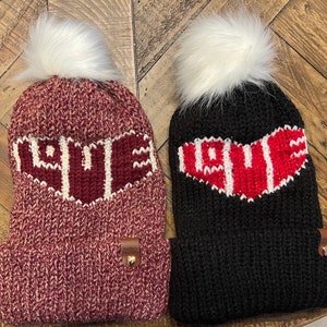 Può includere: Due berretti invernali di lana con pon pon. Un berretto è bordeaux con la scritta "LOVE" in bianco e l'altro è nero con un cuore rosso e la scritta "LOVE" in bianco.