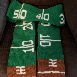 May include: A green and brown knitted scarf with white numbers and lines. The numbers on the scarf are 50, 40, 30, 20, 10, and 0.