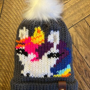 Può includere: Berretto di lana grigio con un pon pon bianco e un design di unicorno colorato. Il berretto ha un'etichetta in pelle marrone con il testo "Goldie's Cozy Knits".