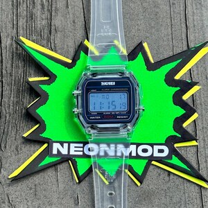 May include: A digital wristwatch with a clear, transparent plastic strap and case. The watch face features a black border with the text SKMEI, ALARM CHRONOGRAPH, and WATER 50M RESIST. The digital display shows the time 11:15 and the date MO 17.
