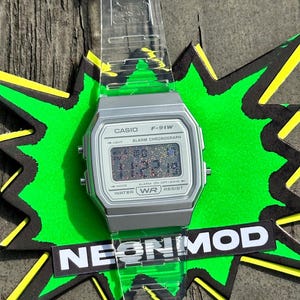 May include: A Casio F-91W digital watch with a silver-toned rectangular case and a clear, transparent resin strap. The watch face features a multi-colored glitter display, black text including CASIO F-91W, and the word NEONMOD on a black label.
