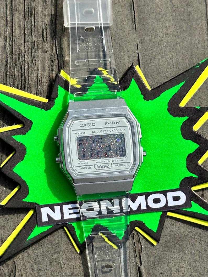 May include: A Casio F-91W digital watch with a silver-toned rectangular case and a clear, transparent resin strap. The watch face features a multi-colored glitter display, black text including CASIO F-91W, and the word NEONMOD on a black label.